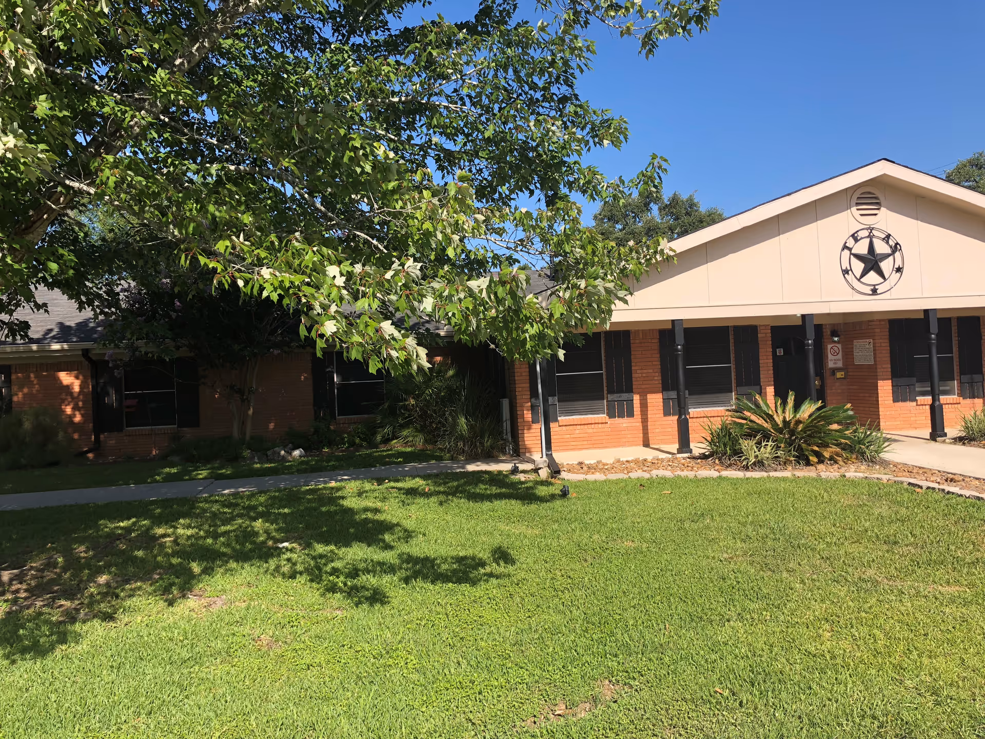 Exterior view of a single-story brick building with a beige gable roof and a large star decoration on the front. The building is surrounded by green grass, shrubs, and a tree with green leaves under a clear blue sky.