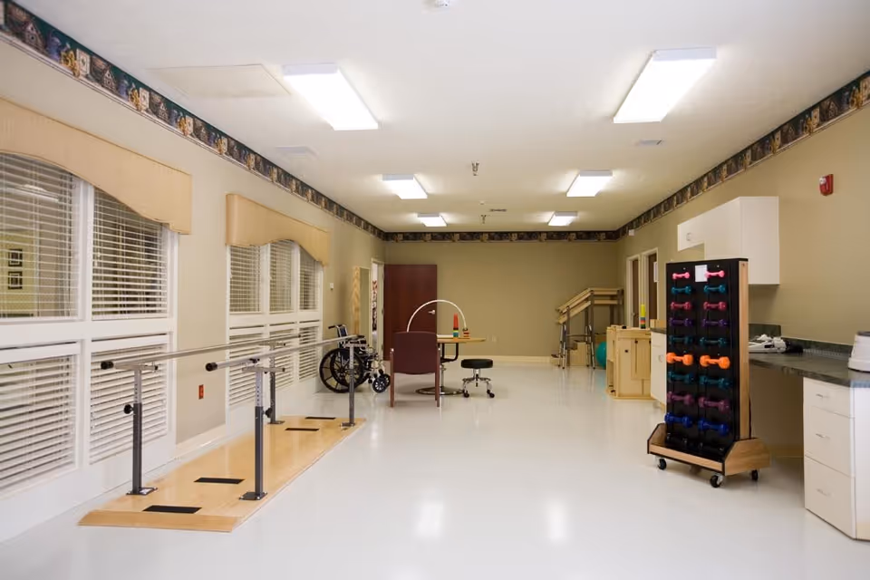 A spacious rehabilitation or physical therapy room with parallel bars for walking exercises, a wheelchair, a table with chairs, and a rack holding colorful dumbbells. The room has large windows with blinds, beige walls, and bright overhead lighting.
