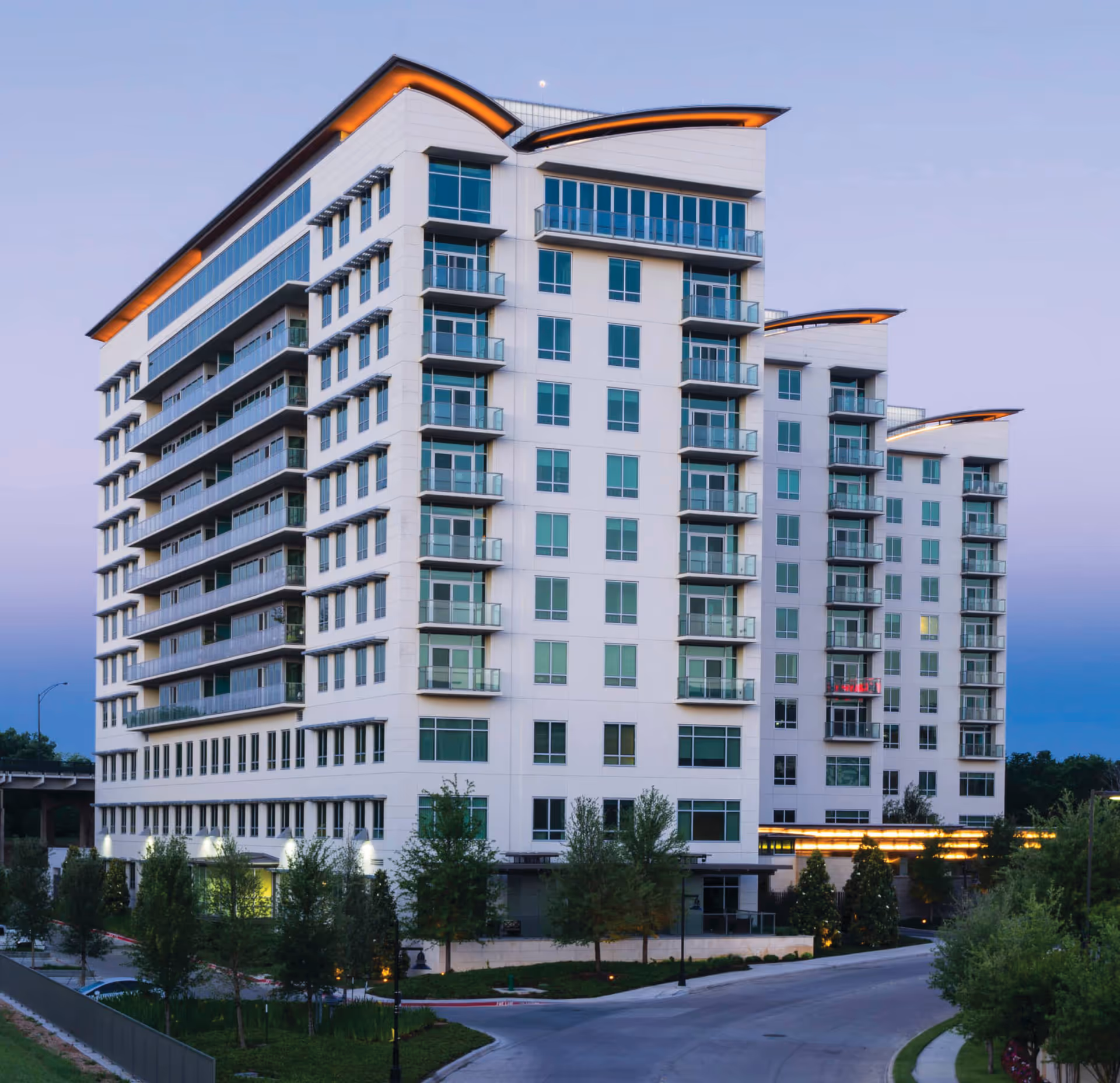 Modern multi-story residential building with balconies and illuminated roof accents, viewed from the front at dusk.