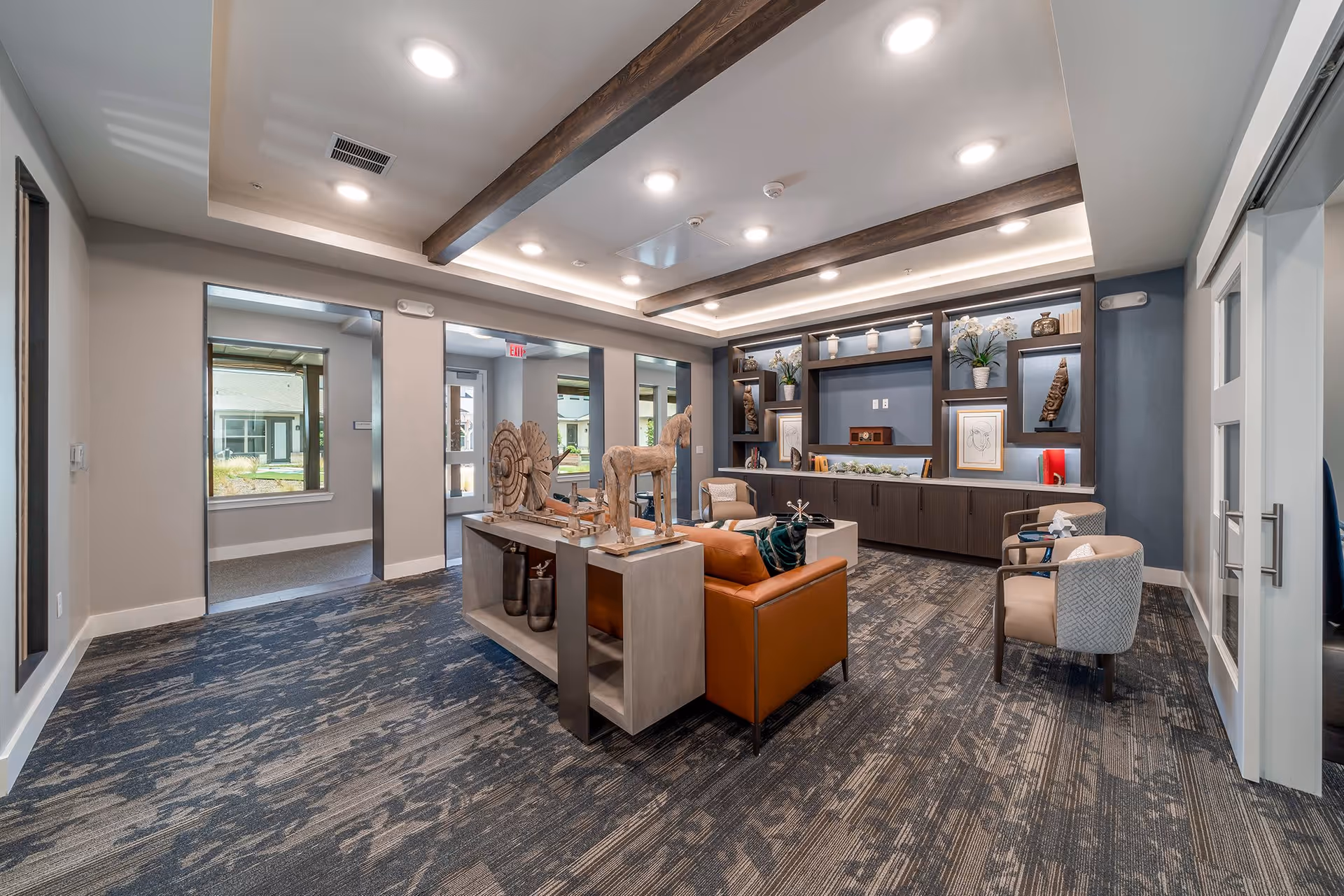 A modern, well-lit common area in a senior living facility with a patterned carpet, wooden ceiling beams, and recessed lighting. The room features a brown leather sofa, two beige armchairs, and a console table with decorative wooden sculptures. Built-in shelves on the far wall display various art pieces, plants, and decorative items. Large windows and glass doors provide natural light and views of the outdoor area.