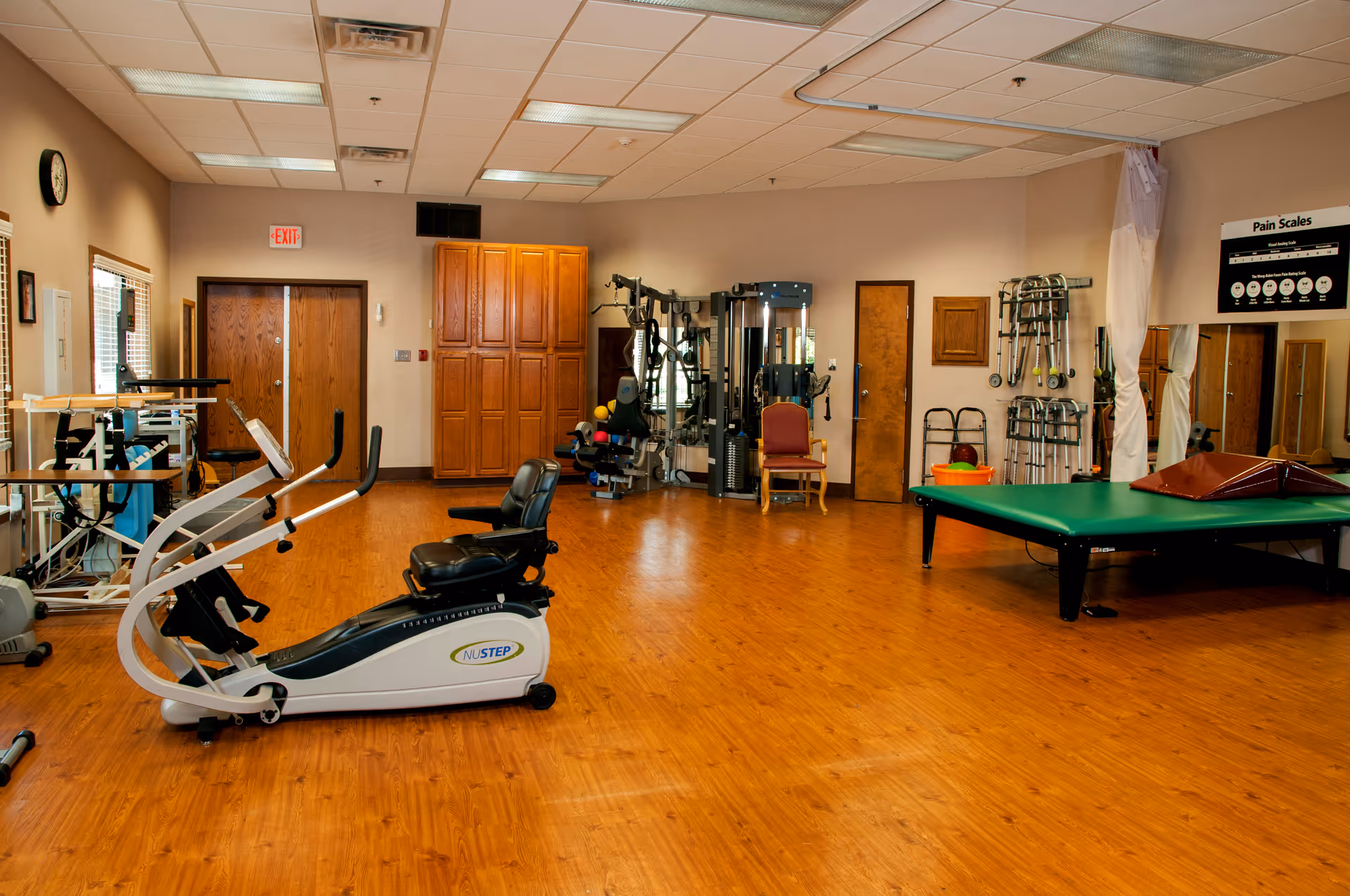 A spacious rehabilitation or physical therapy room with exercise equipment including a NuStep recumbent cross trainer, weight machines, parallel bars, and a large padded therapy table. The room has wooden flooring, beige walls, ceiling lights, and a wall-mounted clock. There are also chairs, a pain scale chart on the wall, and various therapy tools and equipment neatly arranged.