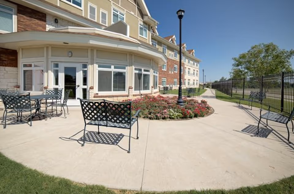 Outdoor patio and landscaped courtyard with metal benches and tables in front of a multi-story senior living building.