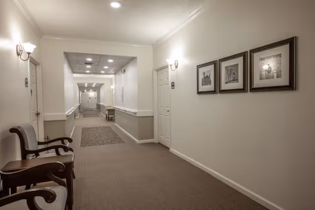 A long, well-lit hallway in a senior living facility with beige walls and carpeted floor. There are two wooden chairs with armrests and a small table on the left side near the foreground. The walls have framed black and white photographs and wall-mounted light fixtures. The hallway extends into the distance with multiple doors and a carpet runner in the middle.