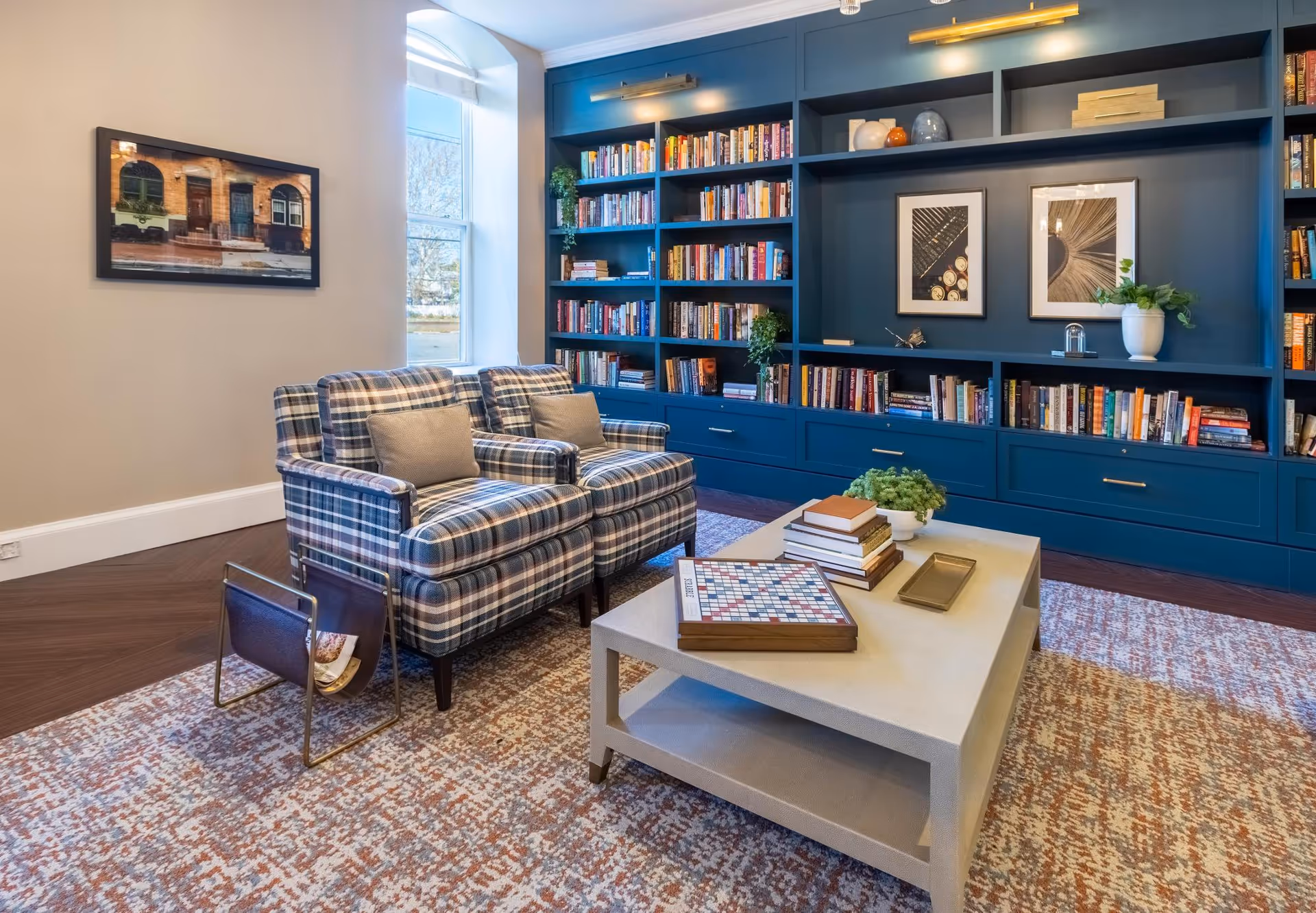 A cozy living room area with two plaid armchairs, a large beige coffee table with books and a plant on it, and a built-in blue bookshelf filled with books and decorative items. There is a large window letting in natural light and a framed picture on the wall.