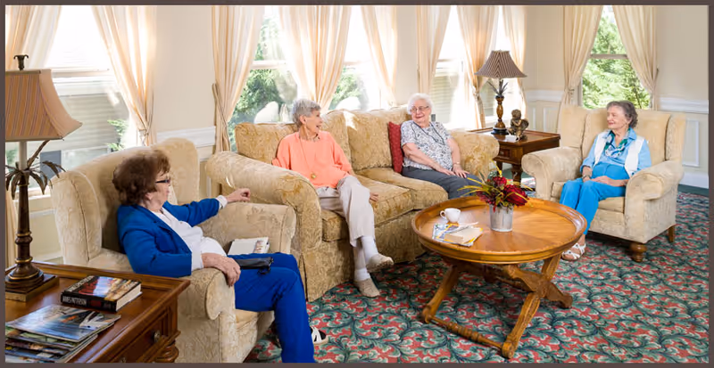 A cozy living room with three elderly women sitting on beige upholstered armchairs and a sofa, engaged in conversation. The room features large windows with sheer curtains, a wooden coffee table with a flower arrangement and books, a patterned carpet, and two table lamps on side tables.