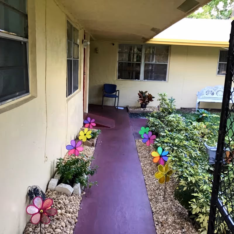 A covered outdoor walkway with a purple floor leading to a door. On either side of the walkway are small garden beds with plants and colorful flower-shaped garden decorations. There is a blue chair near the door and a white bench in the garden area to the right.