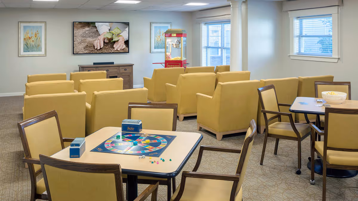 A cozy common area with multiple yellow armchairs arranged in rows facing a flat-screen TV mounted on the wall. There is a popcorn machine near the window. In the foreground, two tables with chairs are set up for playing board games, with one table featuring a Trivial Pursuit game. The room has carpeted floors, light-colored walls, and several windows letting in natural light.