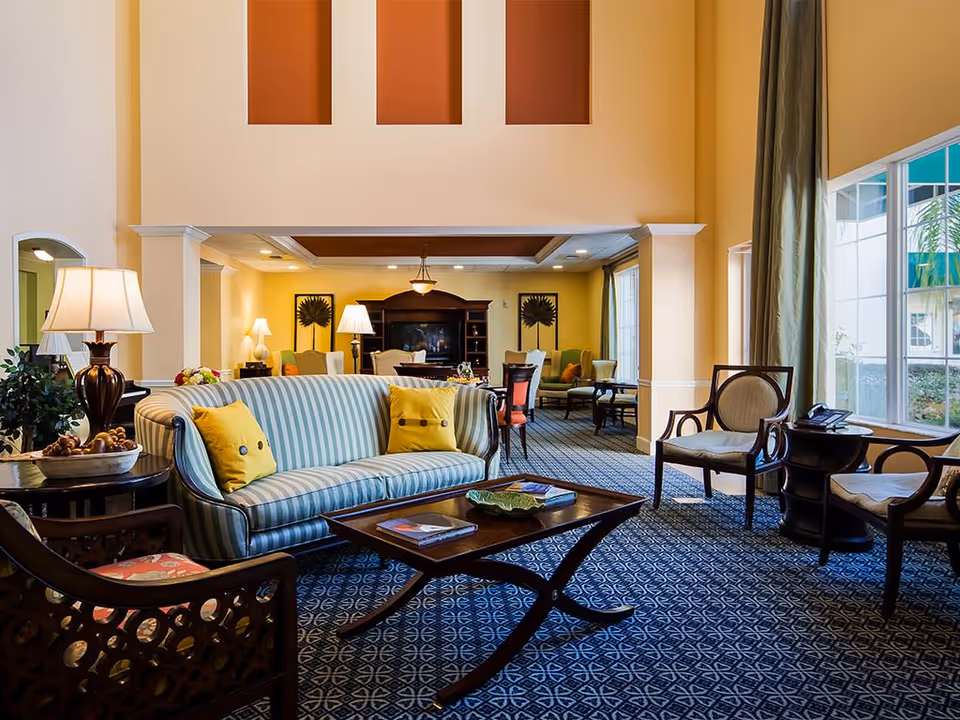 Sunny communal living room with a striped sofa, armchairs, coffee table, lamps and large windows.
