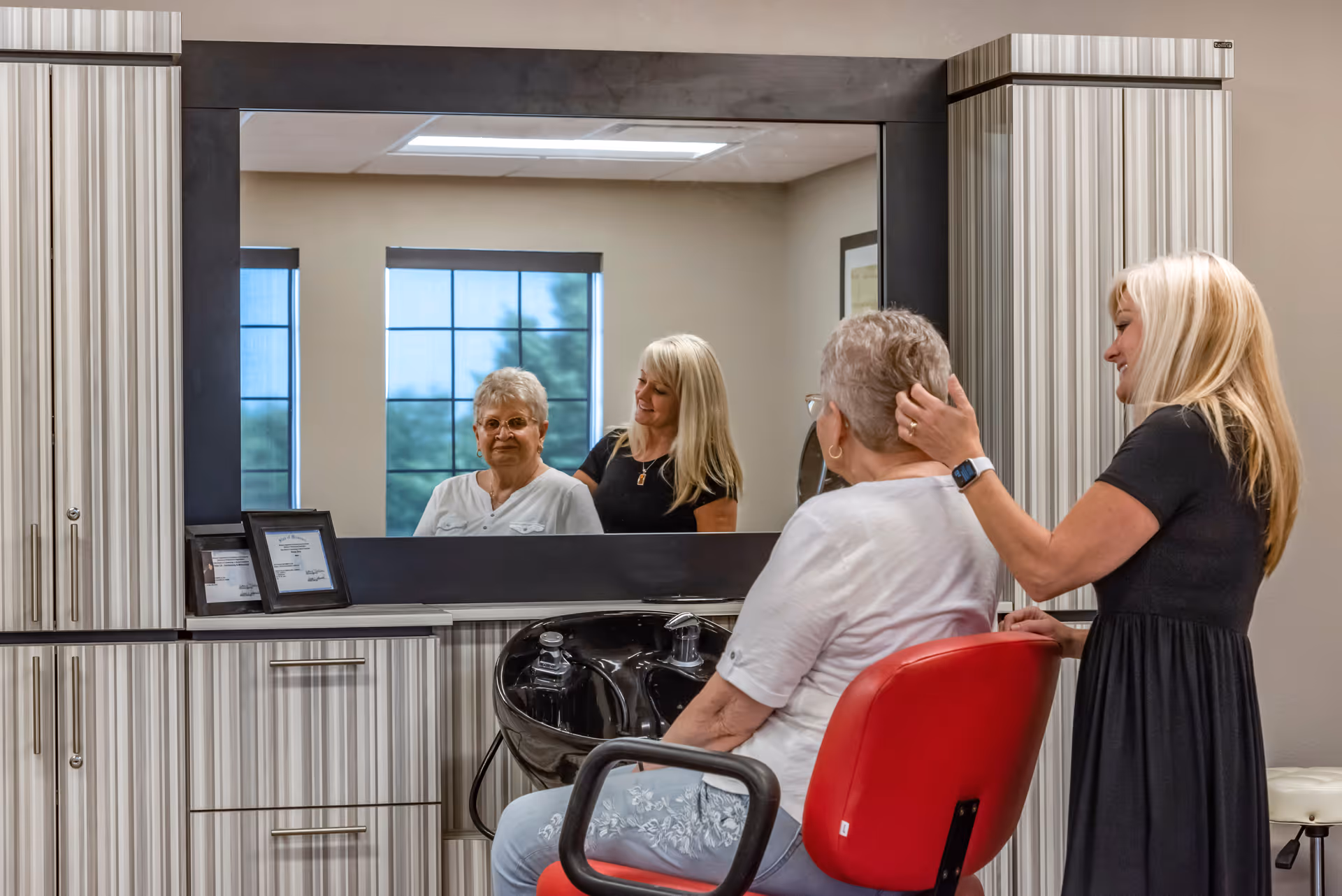 An elderly woman with short gray hair sits in a red salon chair facing a large mirror, while a blonde woman in a black dress stands behind her, gently touching her hair. The room has striped cabinetry and a black sink for hair washing beneath the mirror. Two windows with blue shades are visible in the reflection.