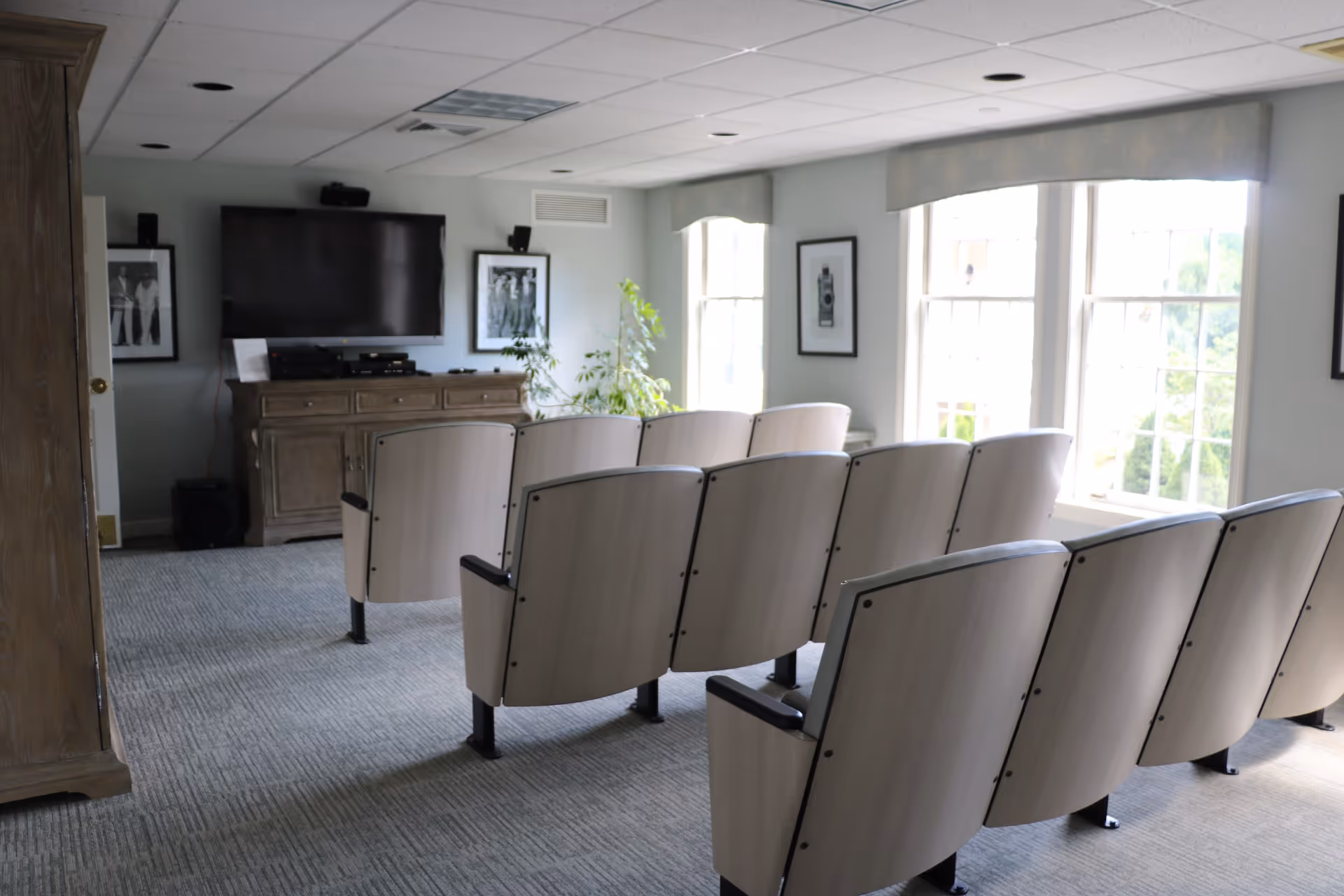 A small theater-style room with two rows of light-colored wooden seats facing a large flat-screen TV mounted on a wall. The room has carpeted flooring, framed black and white photographs on the walls, large windows with valances letting in natural light, and a wooden cabinet beneath the TV with electronic equipment on top.