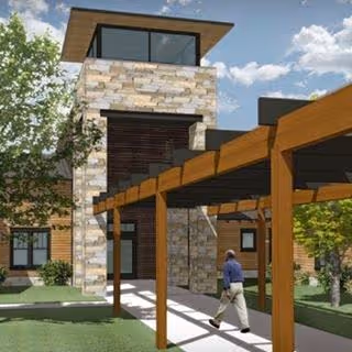 A modern building exterior with a stone tower, wooden pergola walkway, and a person walking along the path.