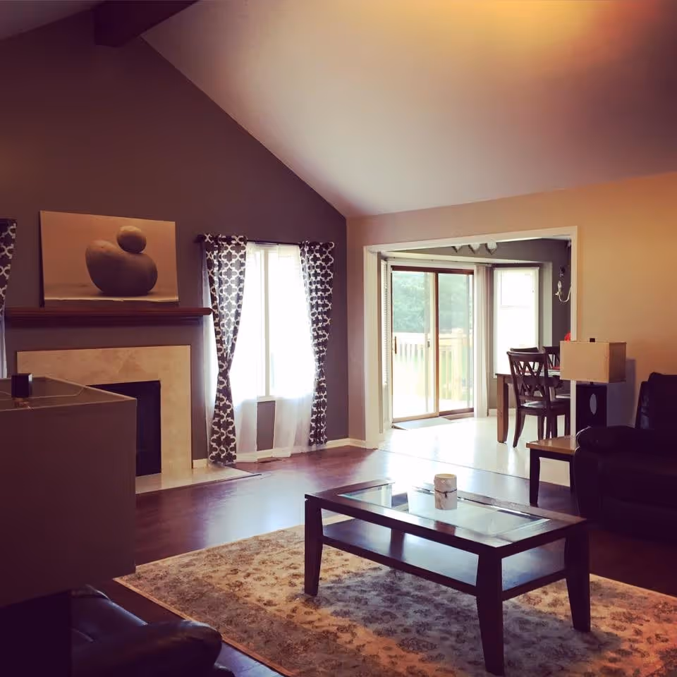 A cozy living room with a high vaulted ceiling, a fireplace with a painting above it, patterned curtains on the window, a glass coffee table on a patterned rug, a leather armchair, and a view into a dining area with sliding glass doors leading outside.