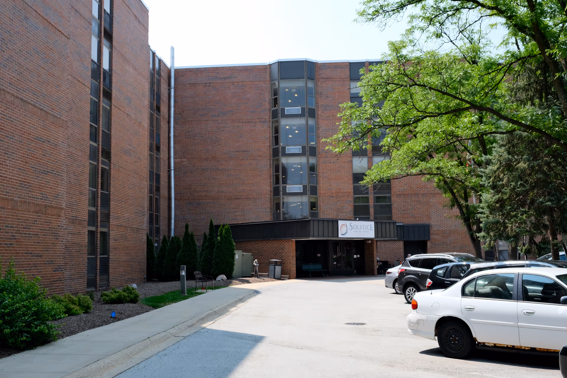 Exterior view of Solstice Senior Living at Palatine, showing a multi-story brick building with large windows and an entrance canopy. Several cars are parked in front, and there are trees and shrubs around the building.