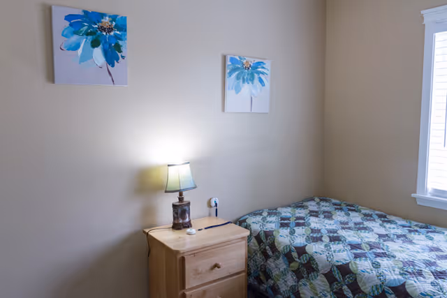 A small bedroom with a single bed covered in a patterned quilt next to a wooden nightstand with a lamp. Two blue floral paintings hang on the beige wall above the nightstand. A window with white blinds is on the right side of the room.