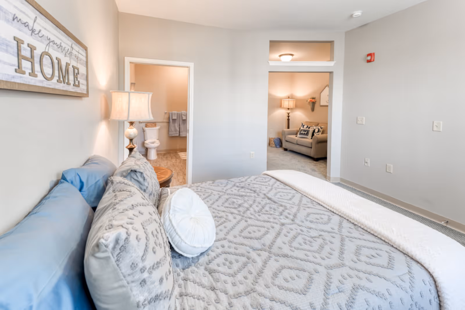 A cozy bedroom with a neatly made bed featuring patterned bedding and multiple pillows. A bedside table with a lamp is next to the bed. On the wall above the bed is a decorative sign that reads 'make yourself at HOME'. Through an open doorway, a bathroom with a toilet and towels is visible. Another open doorway leads to a living area with a sofa, lamp, and wall decor.