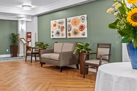 A cozy interior seating area with a gray loveseat and two wooden chairs with cushions, placed against a green wall adorned with two abstract art paintings. There are potted plants beside the furniture and a round table with a white tablecloth and a blue vase holding yellow flowers in the foreground. The floor has a wood pattern, and the space is softly lit with ceiling lights.