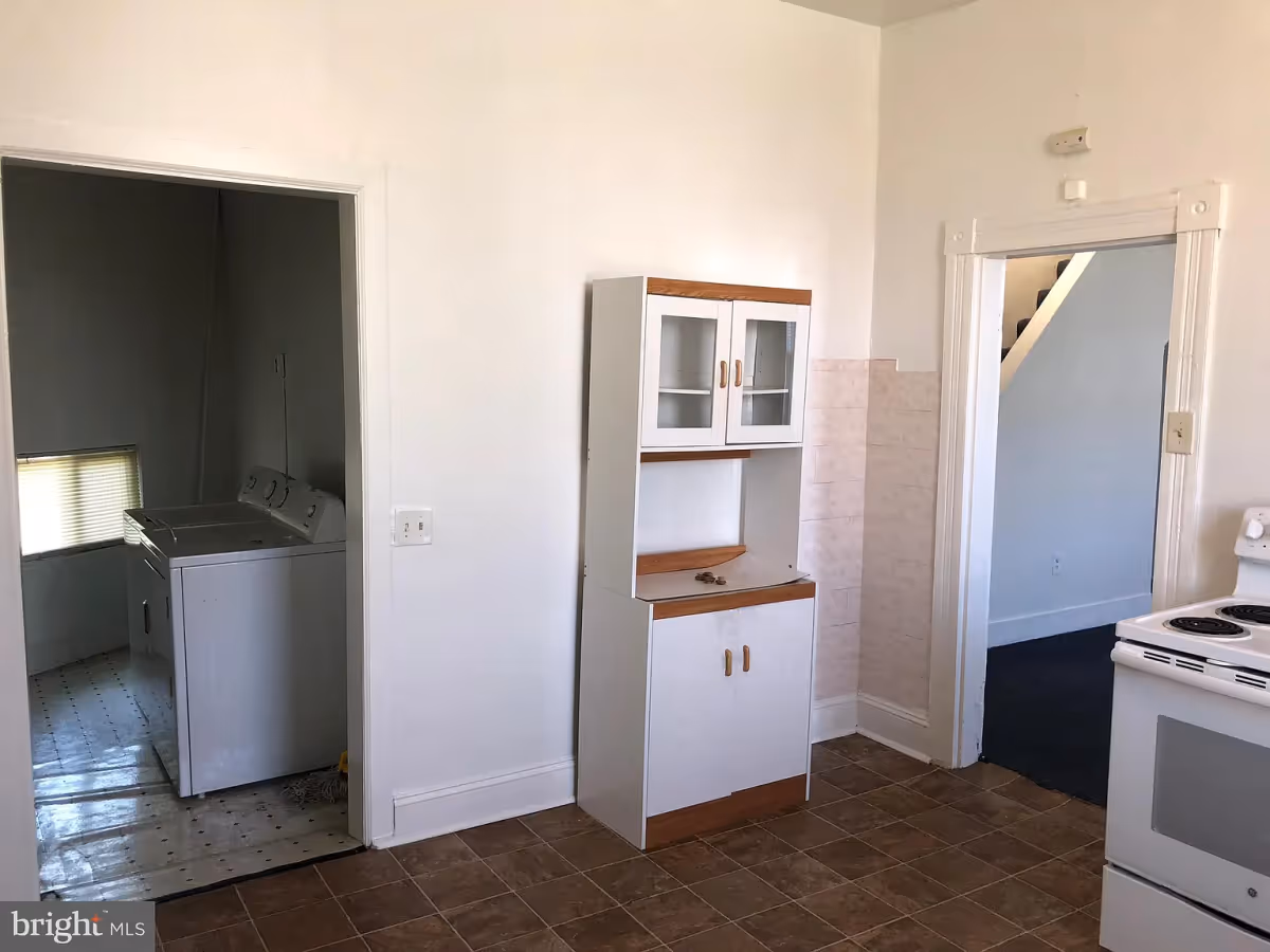 Interior view of a kitchen area with a white stove on the right, a white cabinet with glass doors in the center, and a doorway leading to a laundry room with a washer and dryer on the left. Another doorway on the right leads to a carpeted room with stairs visible.