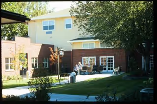 Outdoor courtyard area of a senior living facility with a brick building, green lawn, trees, and a few people sitting on benches near the building entrance.