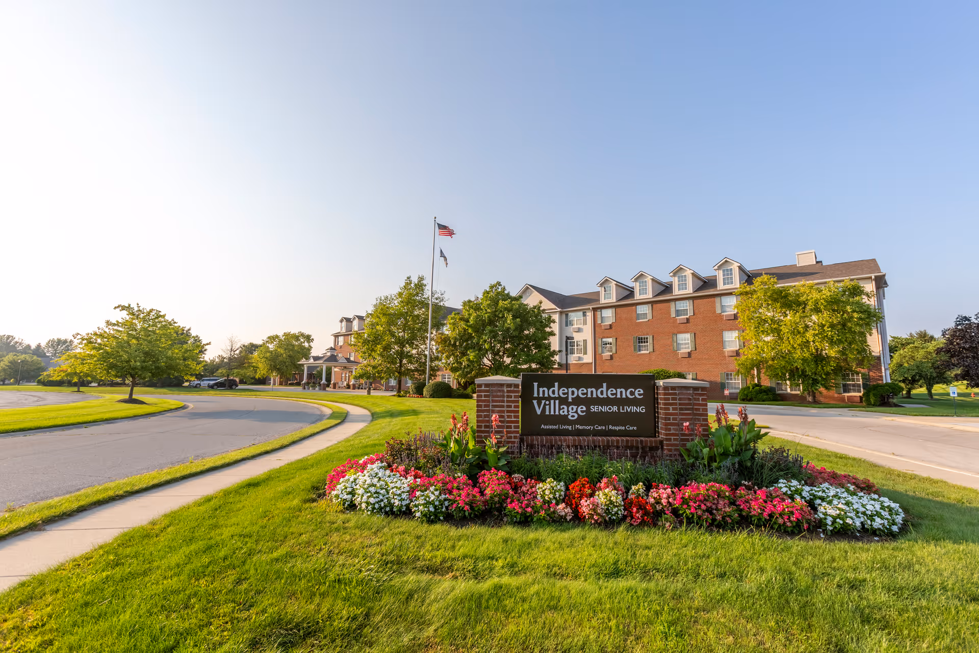 Exterior view of Independence Village Senior Living facility with a brick building, green lawn, colorful flower bed, and two flagpoles with American and state flags under a clear blue sky.