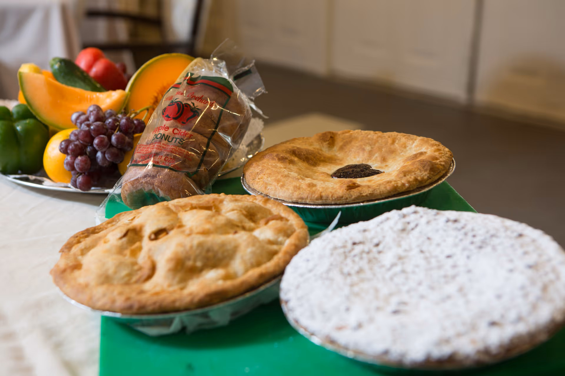 A table with three baked pies, a package of apple cider donuts, and a plate of fresh fruits including grapes, cantaloupe slices, and bell peppers.