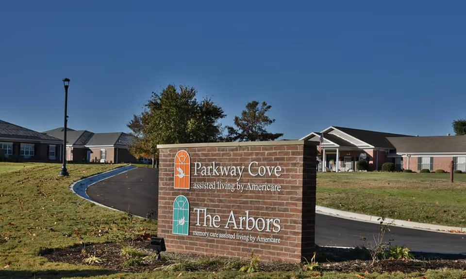 Brick sign at the entrance of a senior living facility with the names Parkway Cove assisted living by Americare and The Arbors memory care assisted living by Americare, with buildings and a driveway in the background under a clear blue sky.