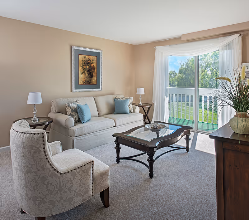 A bright living room with a beige sofa adorned with blue and patterned cushions, a patterned armchair, a glass-top wooden coffee table, two side tables each with a lamp, a framed floral painting on the wall, and a large sliding glass door with sheer white curtains opening to a green outdoor area.