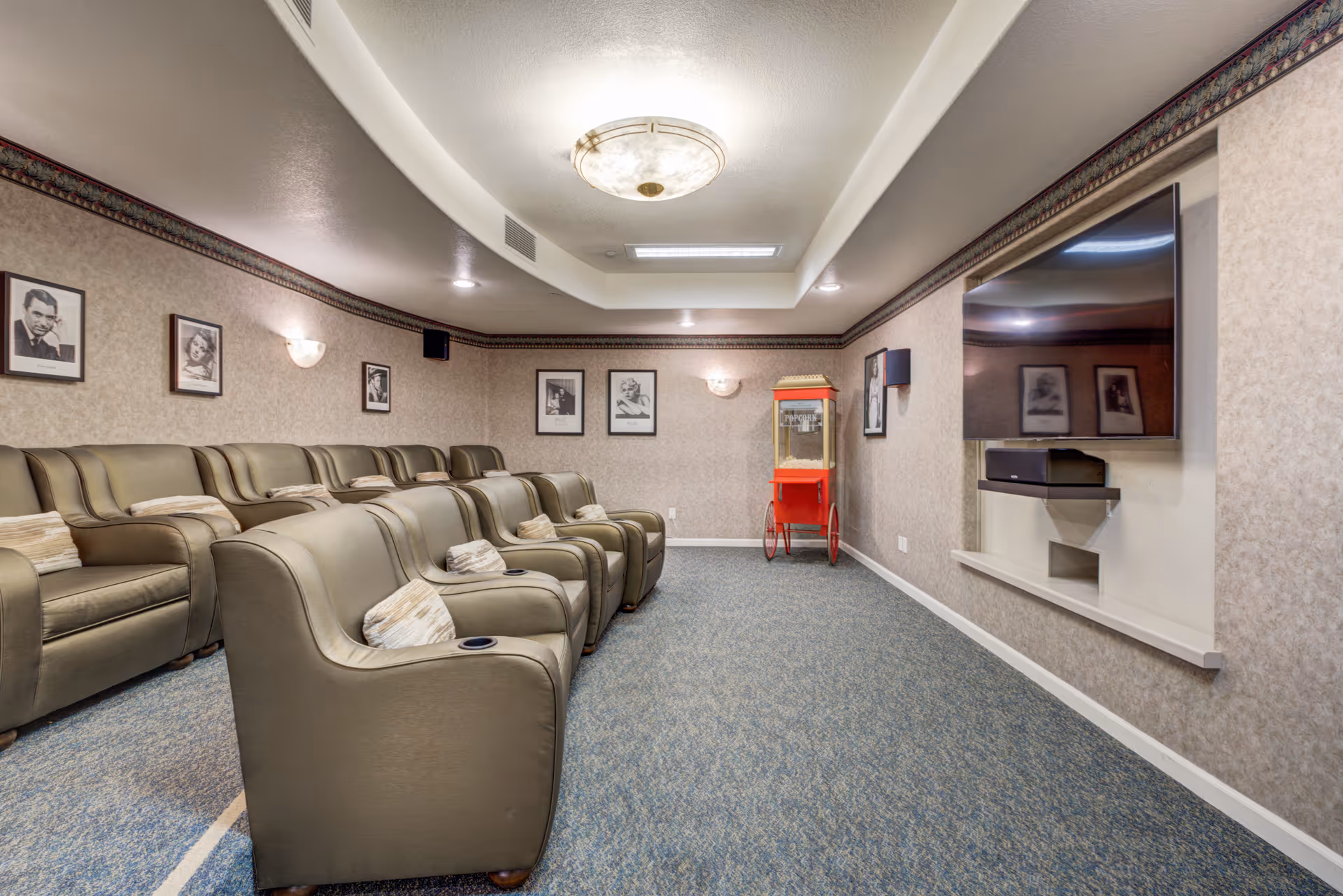 A cozy home theater room with two rows of comfortable leather recliner chairs, each with a small pillow. The walls are decorated with framed black and white portraits, and there is a red popcorn machine in the corner. A large flat-screen TV is mounted on the wall opposite the seating area. The room has soft lighting and carpeted flooring.