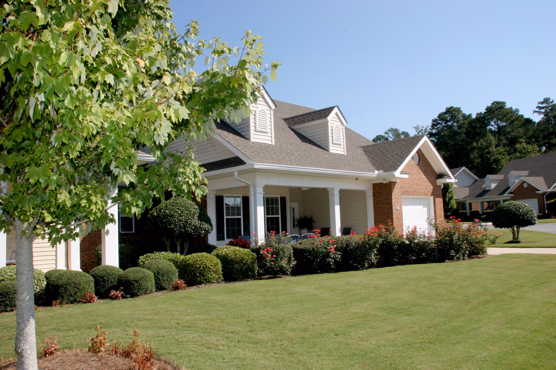 Exterior view of a residential-style building with a well-maintained lawn, bushes, and a tree in the foreground under a clear blue sky.