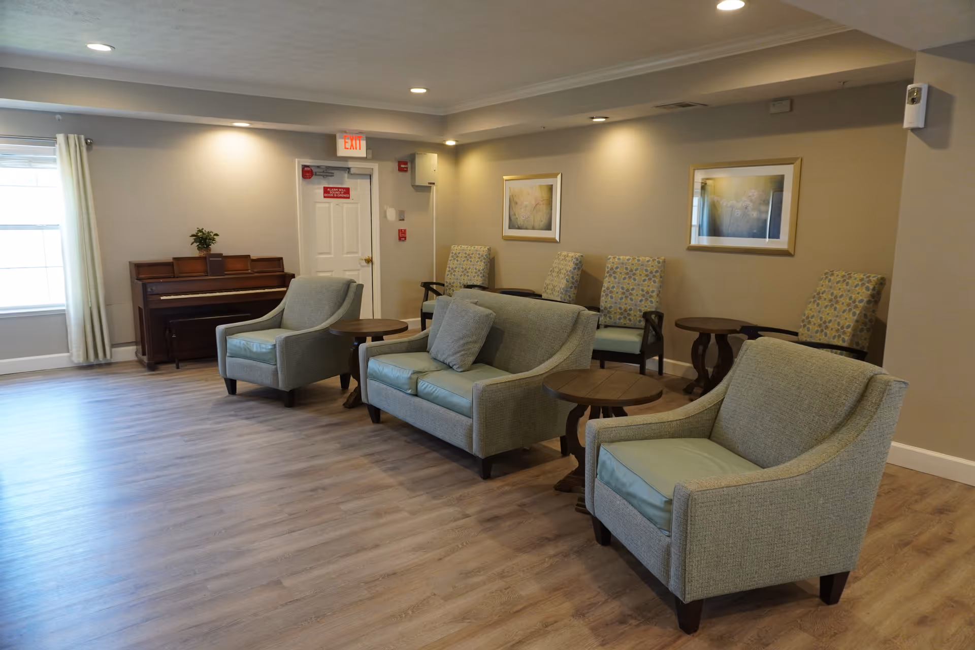 A well-lit living room area with light wood flooring, featuring a piano against the wall near a window with curtains. The room contains a sofa and two armchairs with light blue cushions, several small round wooden tables, and four patterned chairs lined up against the far wall. Two framed pictures hang on the beige walls, and there is an exit door with a red sign above it.