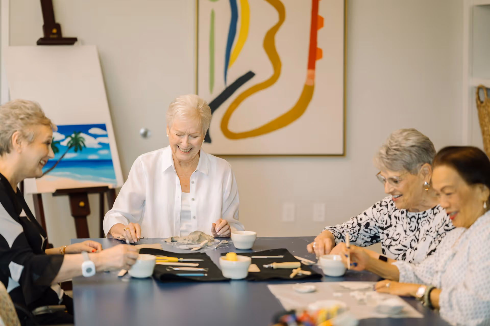 Four elderly women sitting around a table engaged in a creative activity, smiling and enjoying each other's company in a well-lit room with abstract artwork and a painting of a beach on an easel in the background.