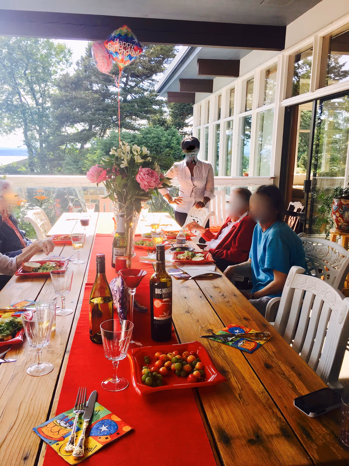 A group of people gathered around a long wooden dining table set with plates of food, wine bottles, glasses, and a vase of flowers. A 'Happy Birthday' balloon is floating above the table. The setting appears to be an outdoor patio with large windows and greenery in the background.