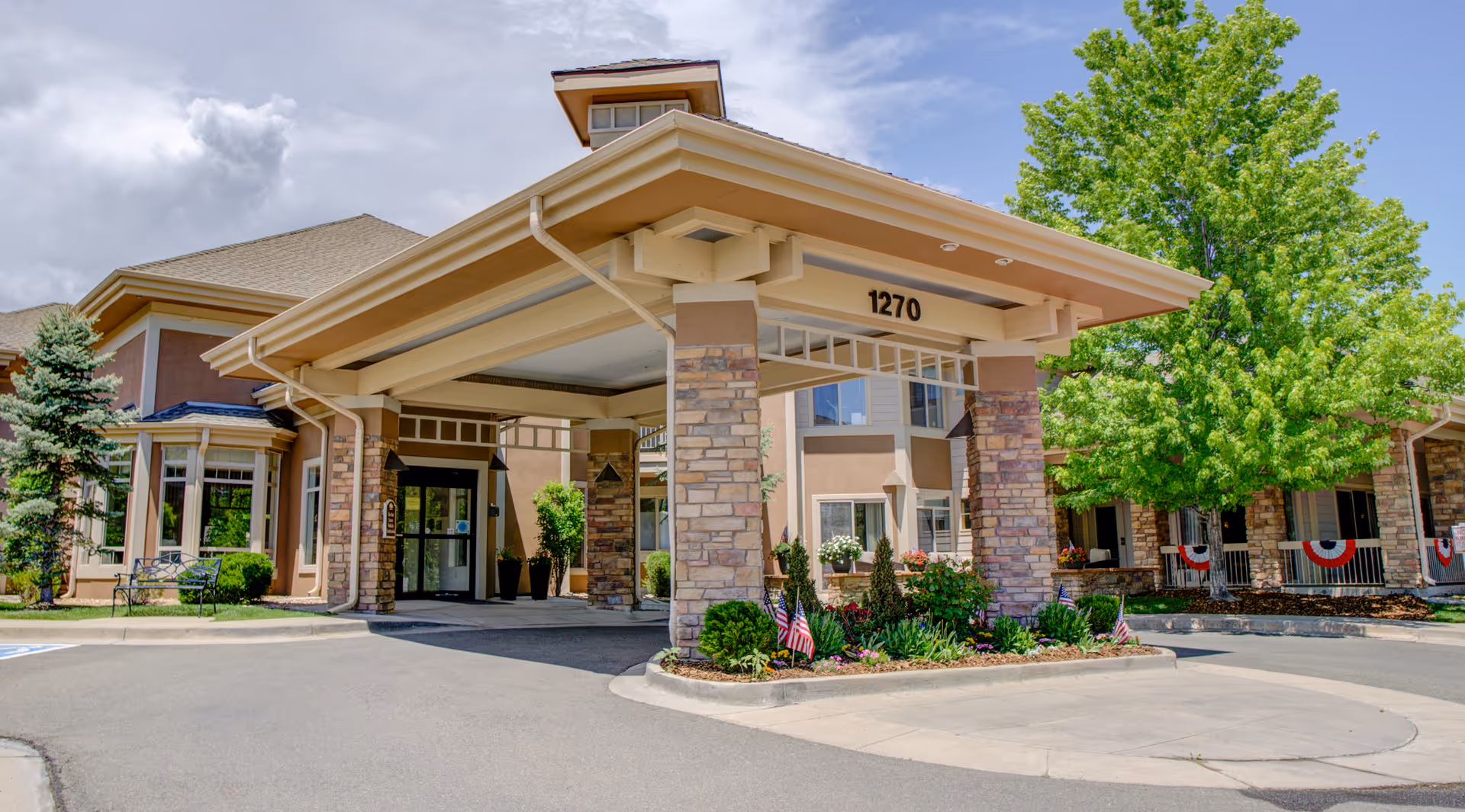 Entrance of a senior living facility with a covered driveway supported by stone pillars, surrounded by well-maintained landscaping including small American flags and green trees under a partly cloudy sky.