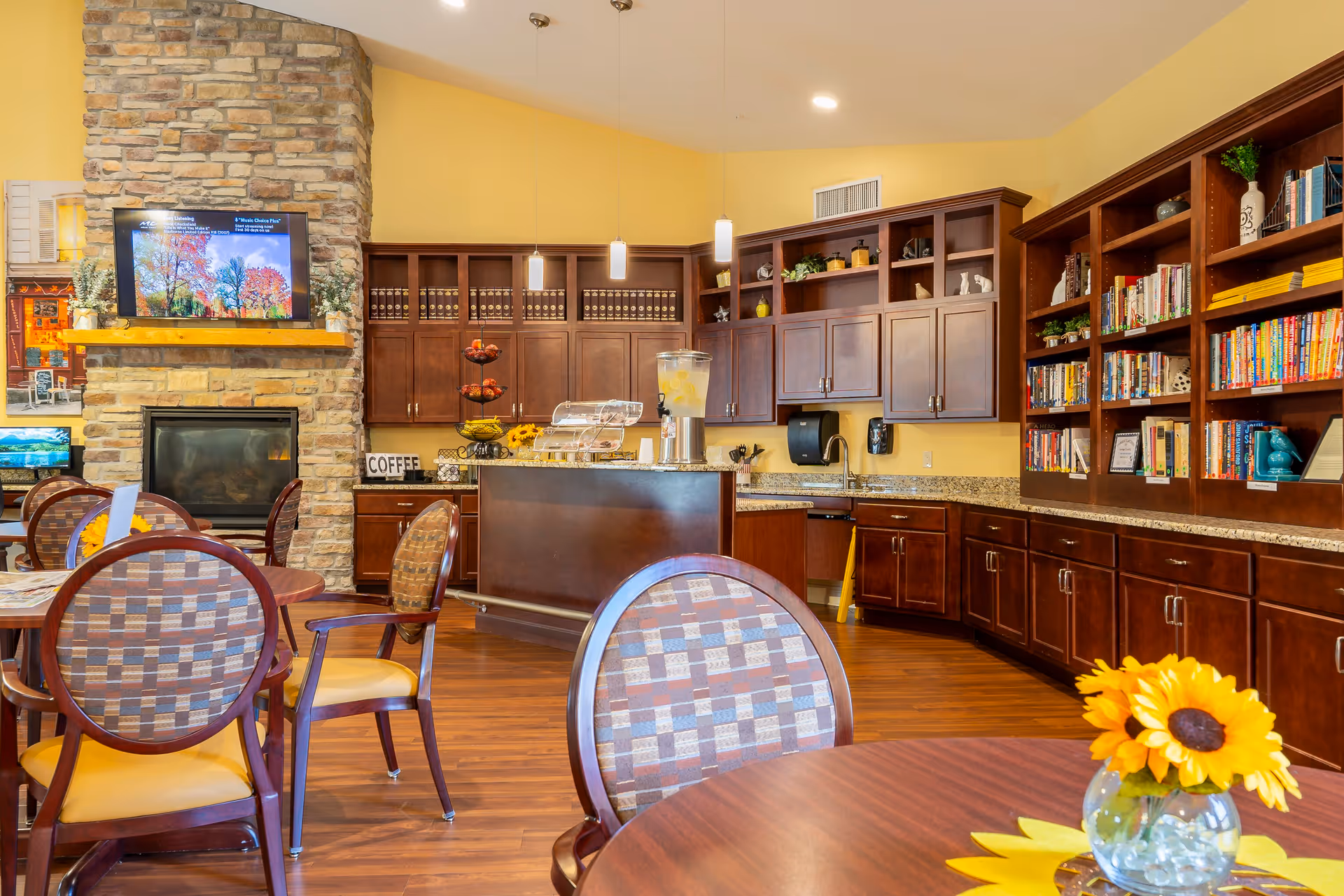 A cozy common area with wooden tables and chairs, a stone fireplace with a mounted TV above it, and a kitchen area with dark wood cabinets and granite countertops. The room has warm yellow walls and wooden flooring. There are bookshelves filled with books and decorative items, a beverage dispenser with lemon water, and a vase with a sunflower on one of the tables.