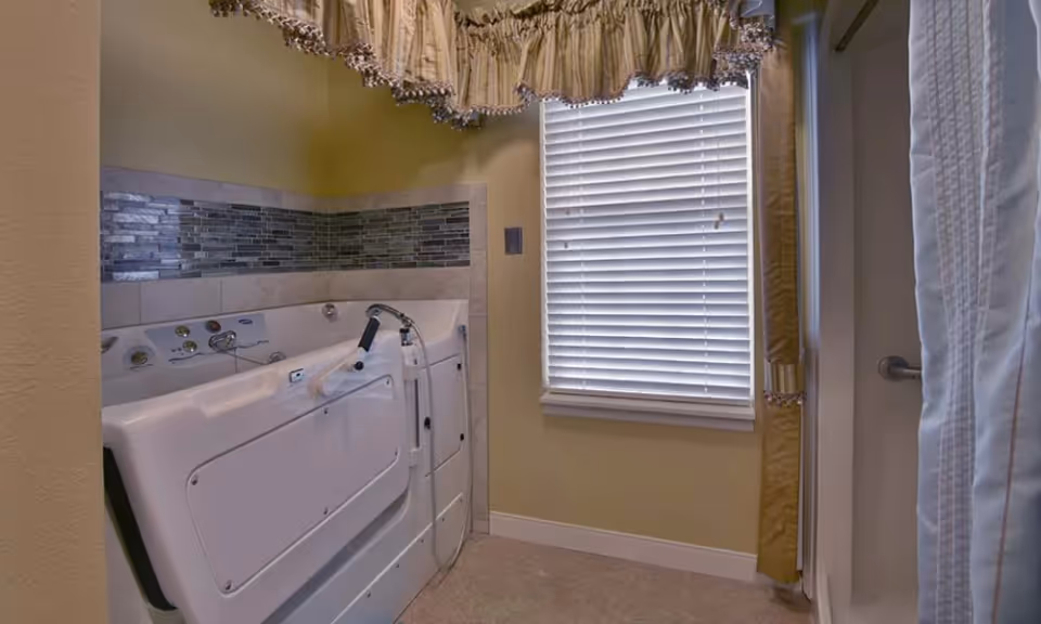 Small bathroom with a white walk-in bathtub, grab bar, tiled backsplash, and a window with blinds and a valance.