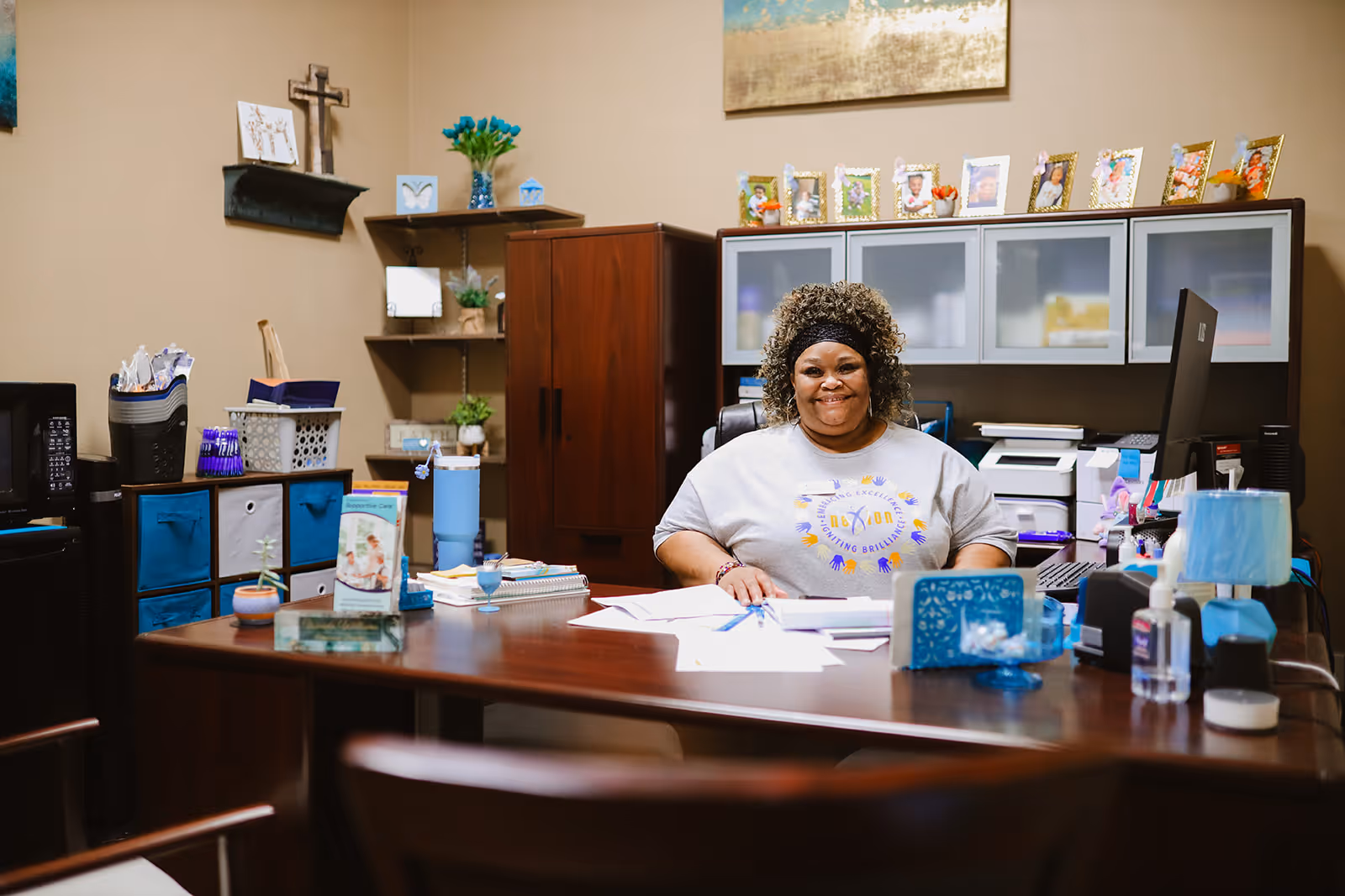 A woman sitting at a desk in an office environment, smiling at the camera. The desk has papers, a computer monitor, a lamp, and various office supplies. Behind her are shelves with framed photos, plants, and office storage units. The room has warm beige walls and a cross hanging on the wall.