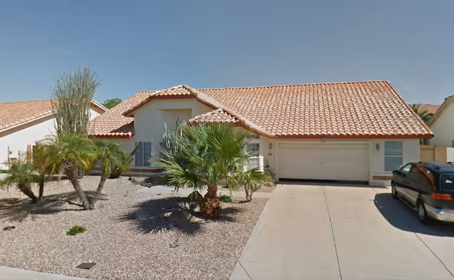 Single-story house with a tiled roof, a two-car garage, and a driveway with a parked minivan. The front yard is landscaped with gravel and several palm trees and desert plants.