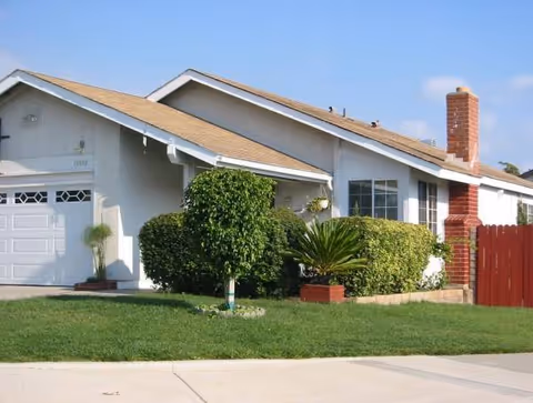 A single-story house exterior with a manicured lawn, shrubs, and a driveway.