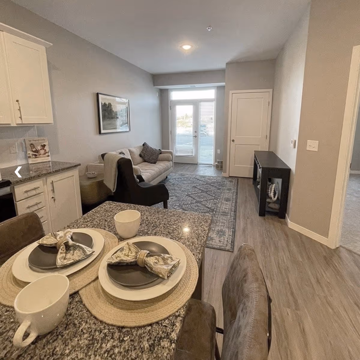 Interior view of a senior living facility showing a combined kitchen and living area. The foreground features a granite countertop with two place settings including plates, napkins, and cups. The living area has a beige sofa with a dark armchair, a patterned rug, a small black console table, and a door with glass panels leading outside. The walls are painted light gray and the flooring is wood-style laminate.
