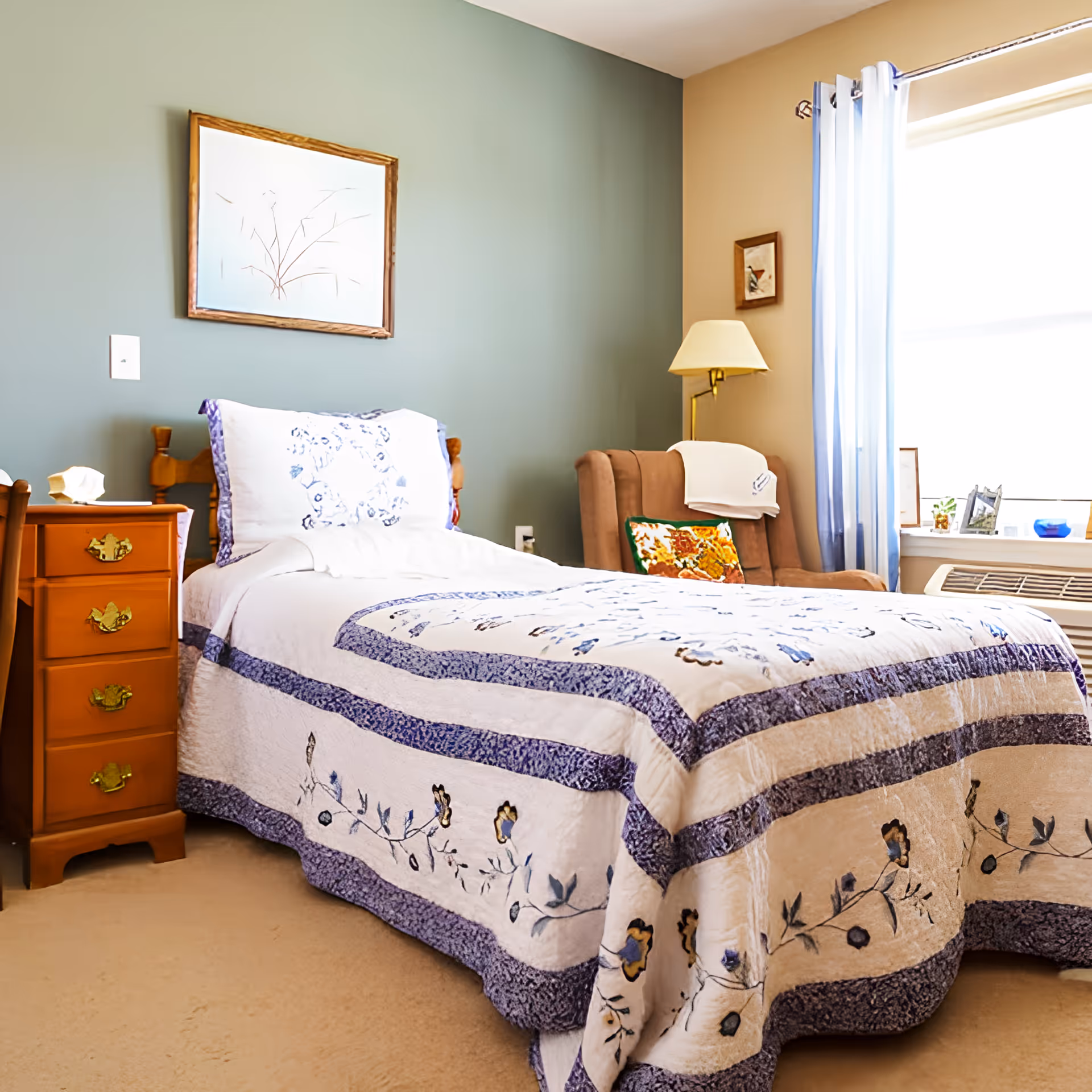 A cozy bedroom with a single bed covered in a white and purple floral quilt. Next to the bed is a wooden nightstand with three drawers and a tissue box on top. A cushioned armchair with a floral pillow and a white cloth draped over the back is positioned near a window with sheer white curtains. A floor lamp and framed artwork are on the walls, which are painted in muted green and beige tones.