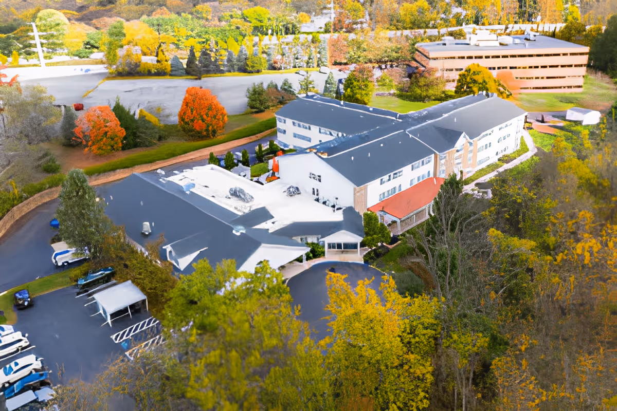 Aerial view of Rittenhouse Village At Lehigh Valley facility surrounded by trees with autumn foliage. The building has a large parking area with several parked vehicles and is situated near another large building in a green, wooded area.