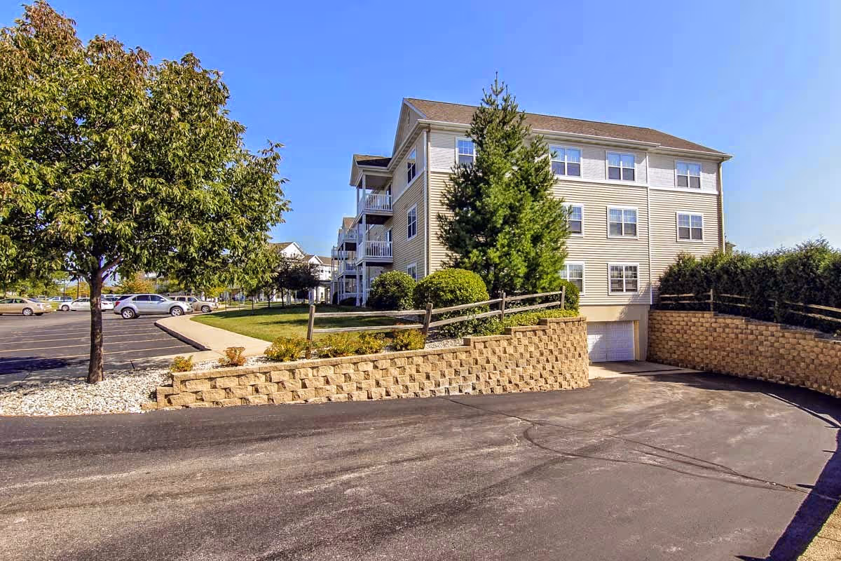 Front exterior of a three-story senior apartment building with a parking area, trees, and a retaining wall.