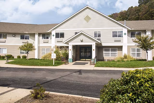Front exterior view of a two-story senior living facility building with beige siding, multiple windows, a central entrance with a covered porch, and landscaped greenery including bushes and small trees.