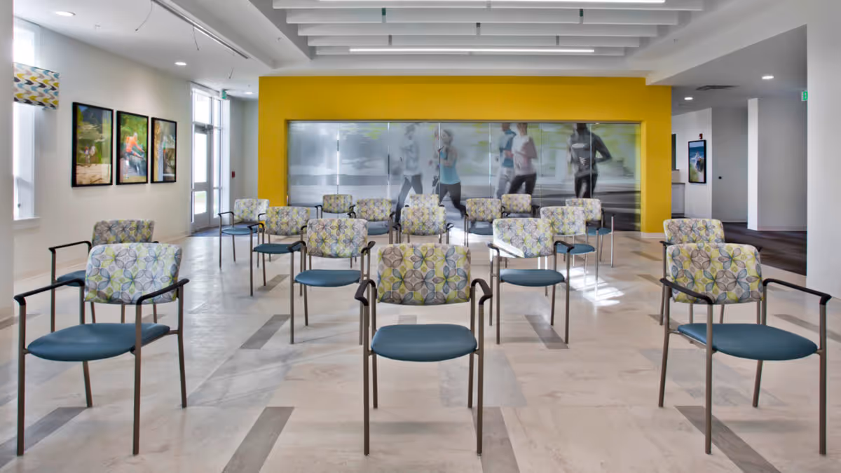 A spacious room with multiple chairs arranged in rows, each chair having a patterned backrest and blue seat. The room features a yellow accent wall with a large glass panel displaying an image of people jogging. There are framed pictures on the left wall and large windows allowing natural light to enter.