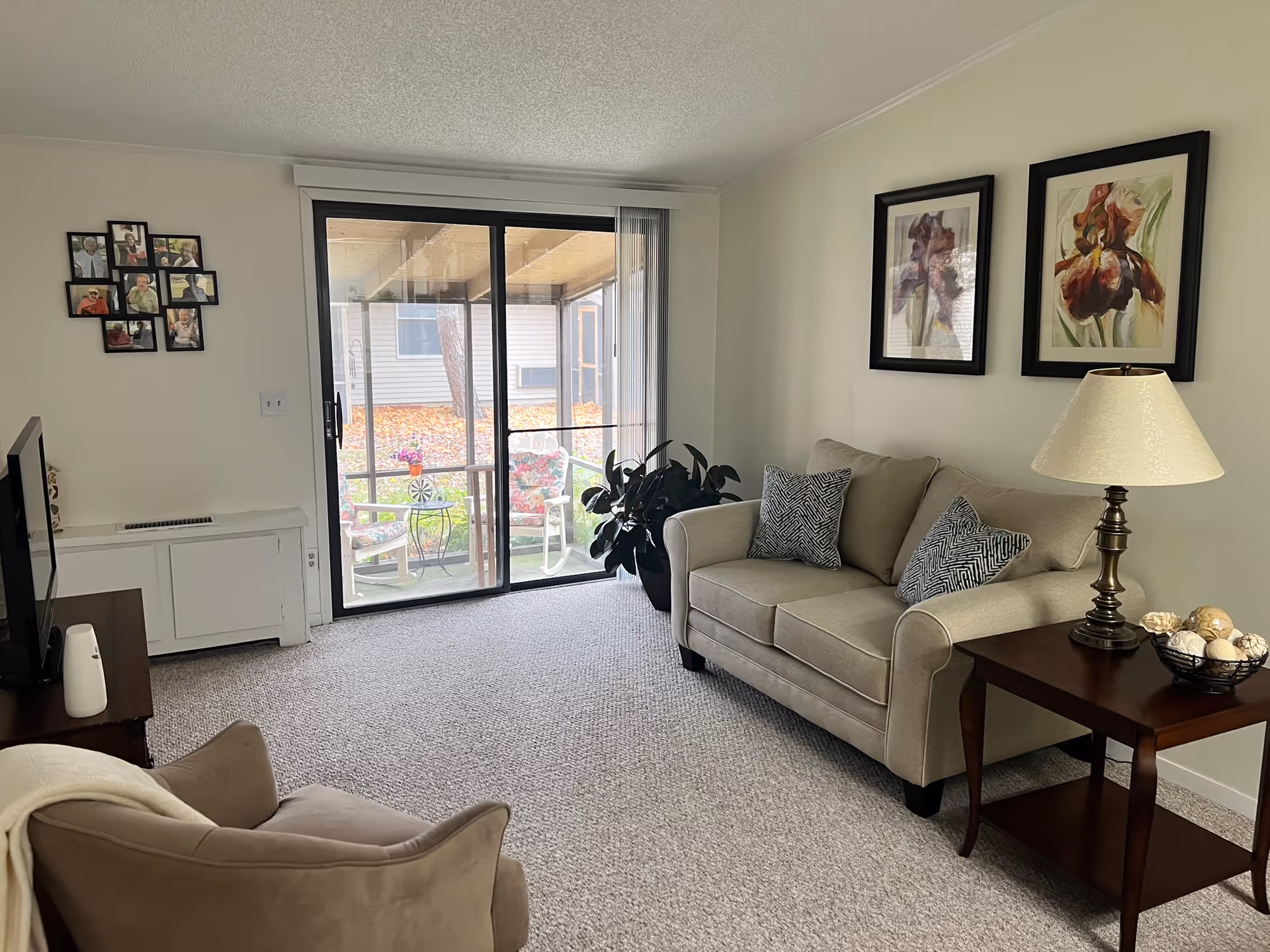 A cozy living room with a beige loveseat adorned with two patterned cushions, a wooden side table with a lamp and decorative bowl, a beige armchair with a blanket draped over it, a TV on a stand, and a sliding glass door leading to a covered patio with floral cushioned chairs. The walls are decorated with framed floral artwork and a collage of photos.