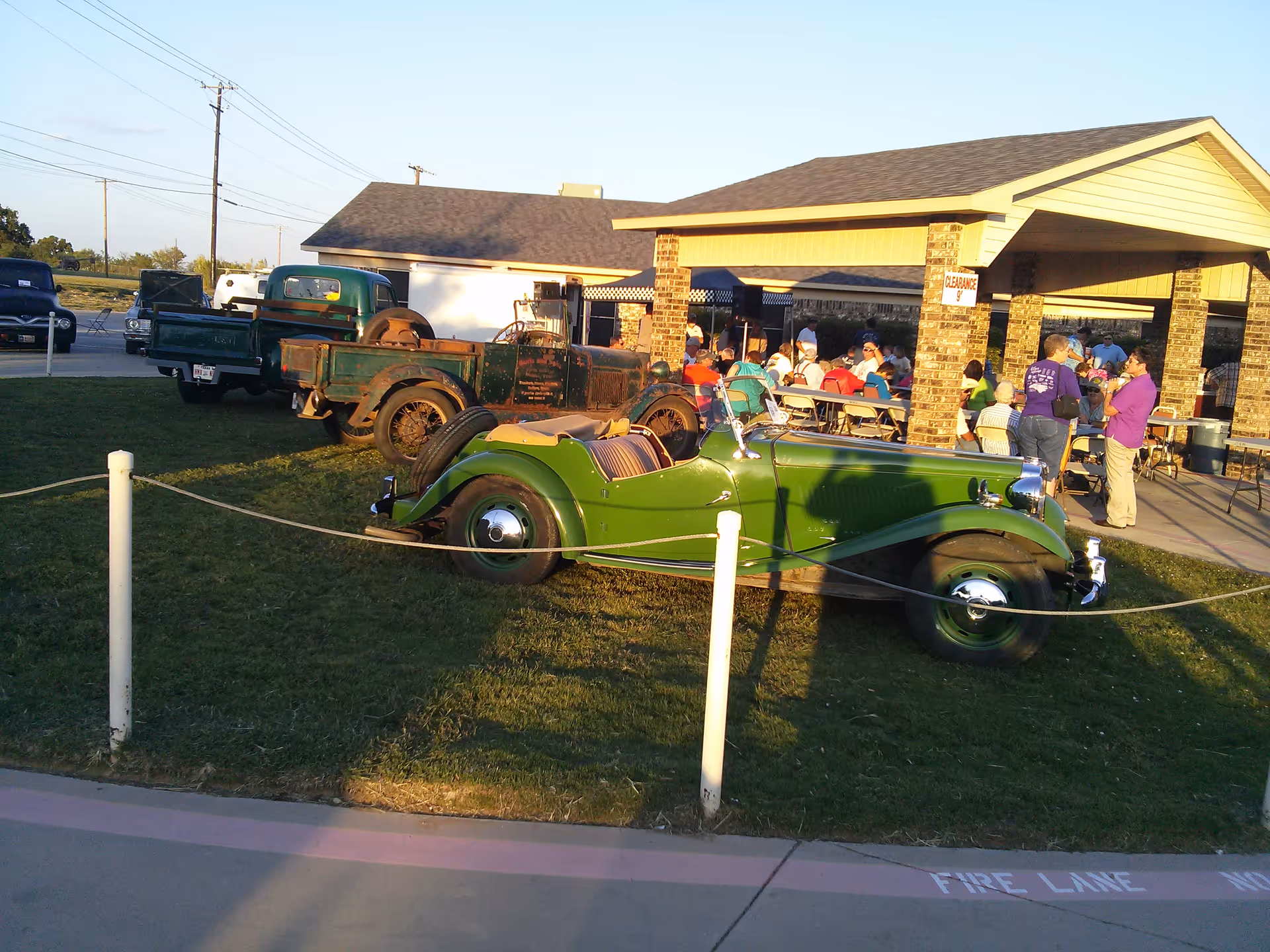 Outdoor scene showing a green vintage convertible car and an old rusty truck parked on grass near a covered pavilion. Several people are seated and standing under the pavilion, which has brick pillars and a roof. The area is roped off and there is a sidewalk with a fire lane marking in the foreground.