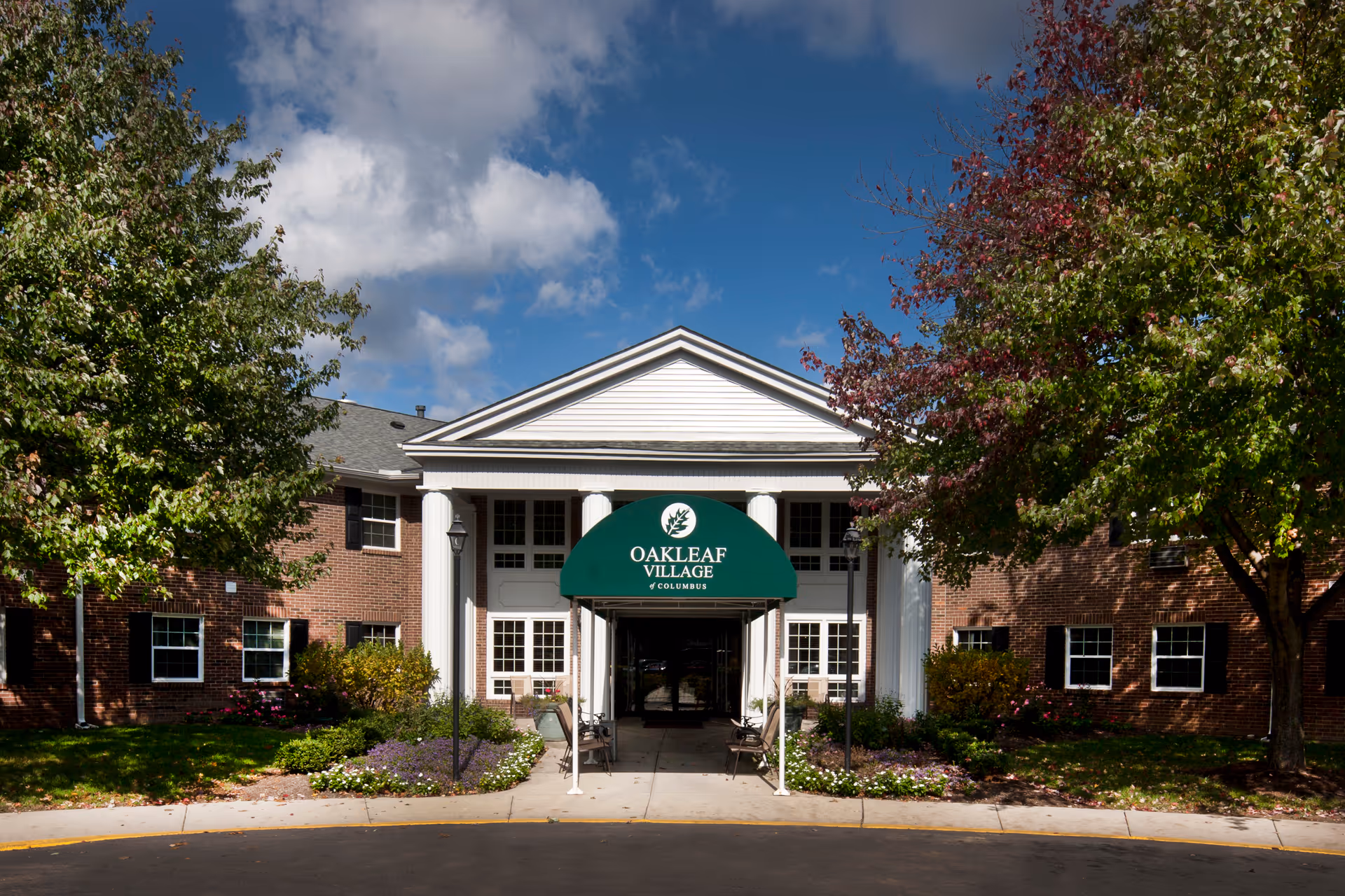 Front exterior view of Oakleaf Village of Columbus, a brick building with white columns and a green awning over the entrance. The building is surrounded by trees and landscaped flower beds under a partly cloudy sky.