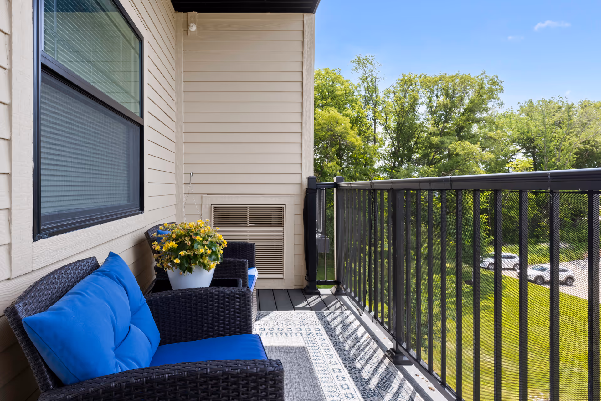 A small balcony with black metal railing overlooking a green area with trees and parked cars. The balcony has two black wicker chairs with blue cushions and a white pot with yellow flowers placed on one of the chairs. The building exterior is beige with horizontal siding and a window with closed blinds.