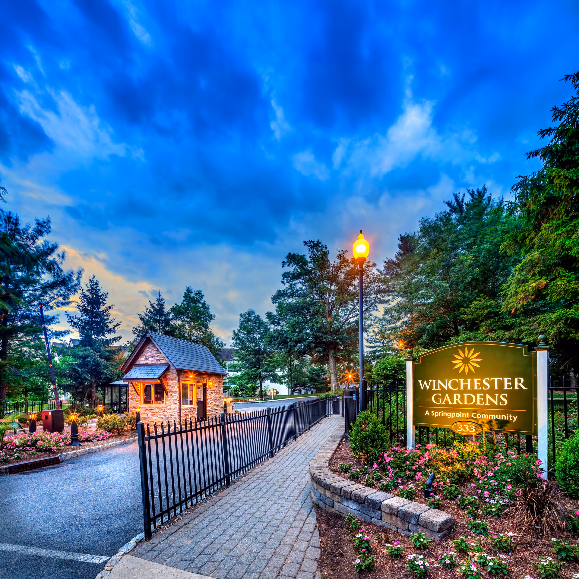 Entrance to Winchester Gardens, a Springpoint Community, featuring a small stone guardhouse, a black metal fence, a lit streetlamp, and a green sign with the community name surrounded by landscaped flowers and trees under a dramatic blue sky at dusk.