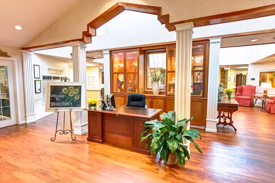 Reception area of Brookstone Assisted Living Community featuring a wooden desk with a computer and chair, a decorative plant, white columns, a chalkboard sign welcoming visitors, wooden flooring, and a seating area with red upholstered chairs in the background.