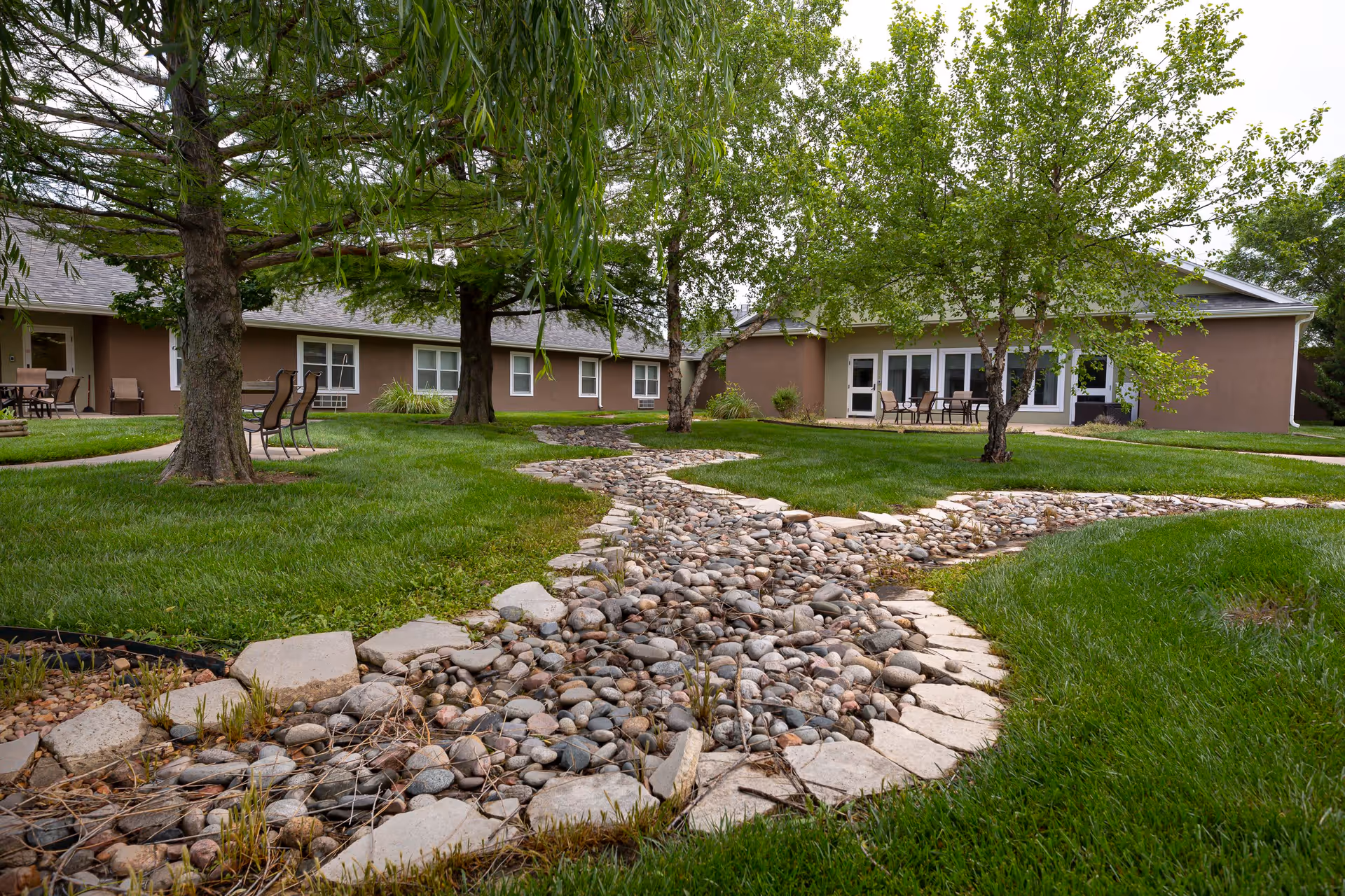 A landscaped outdoor area at Homestead Assisted Living & Memory Care of Wichita featuring a dry creek bed lined with stones and bordered by grass. Several trees provide shade, and there are patio tables and chairs near the building in the background.